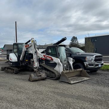 A white Bobcat excavator and a white track loader parked next to a black pickup truck on a gravel lot under a cloudy sky.