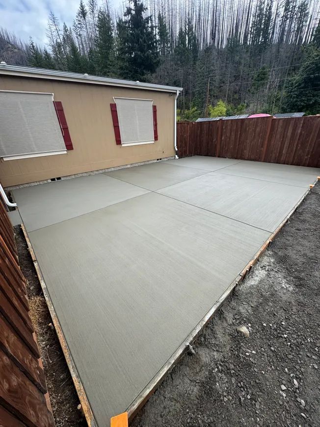A newly poured, rectangular concrete patio adjacent to a beige house with two windows and a wooden fence.