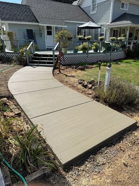A new, smooth concrete walkway leads to the wooden steps of a house deck on a sunny day.