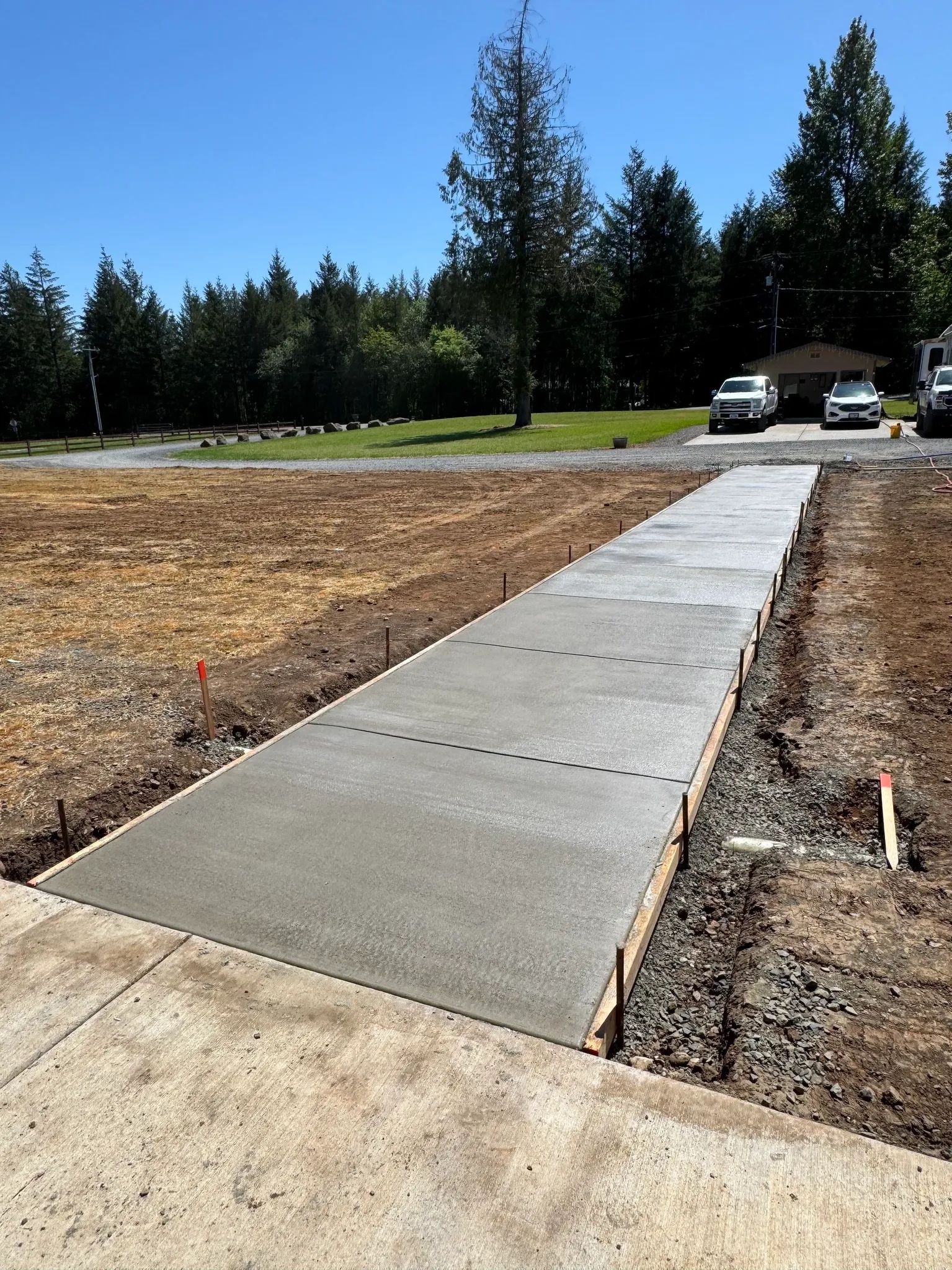 A newly poured concrete walkway extends through a gravel yard toward a driveway with parked cars on a sunny day.