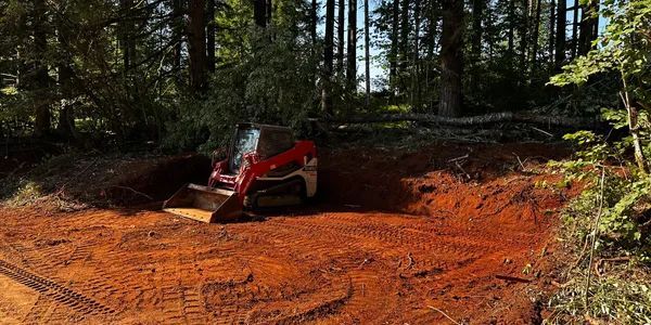 A red skid steer loader parked in a clearing of orange dirt within a forest.