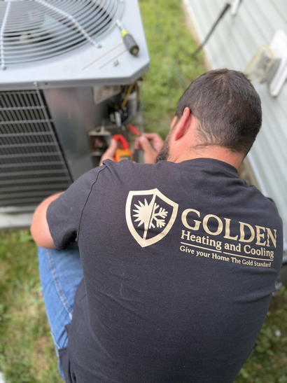 Technician kneels beside an air conditioner, wearing a black Golden Heating and Cooling Services shirt.