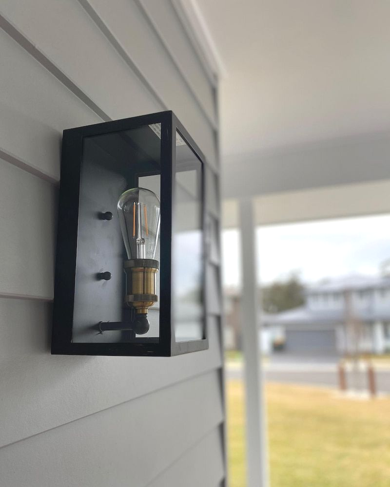 A modern rectangular black metal outdoor wall lantern with an exposed vintage-style Edison bulb on a light grey siding — Hspark Electrical PTY LTD in Austinmer, NSW