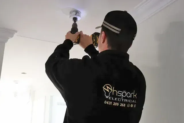An electrician in a black uniform uses a hole saw drill to cut into a white ceiling — Hspark Electrical PTY LTD in Woonona, NSW