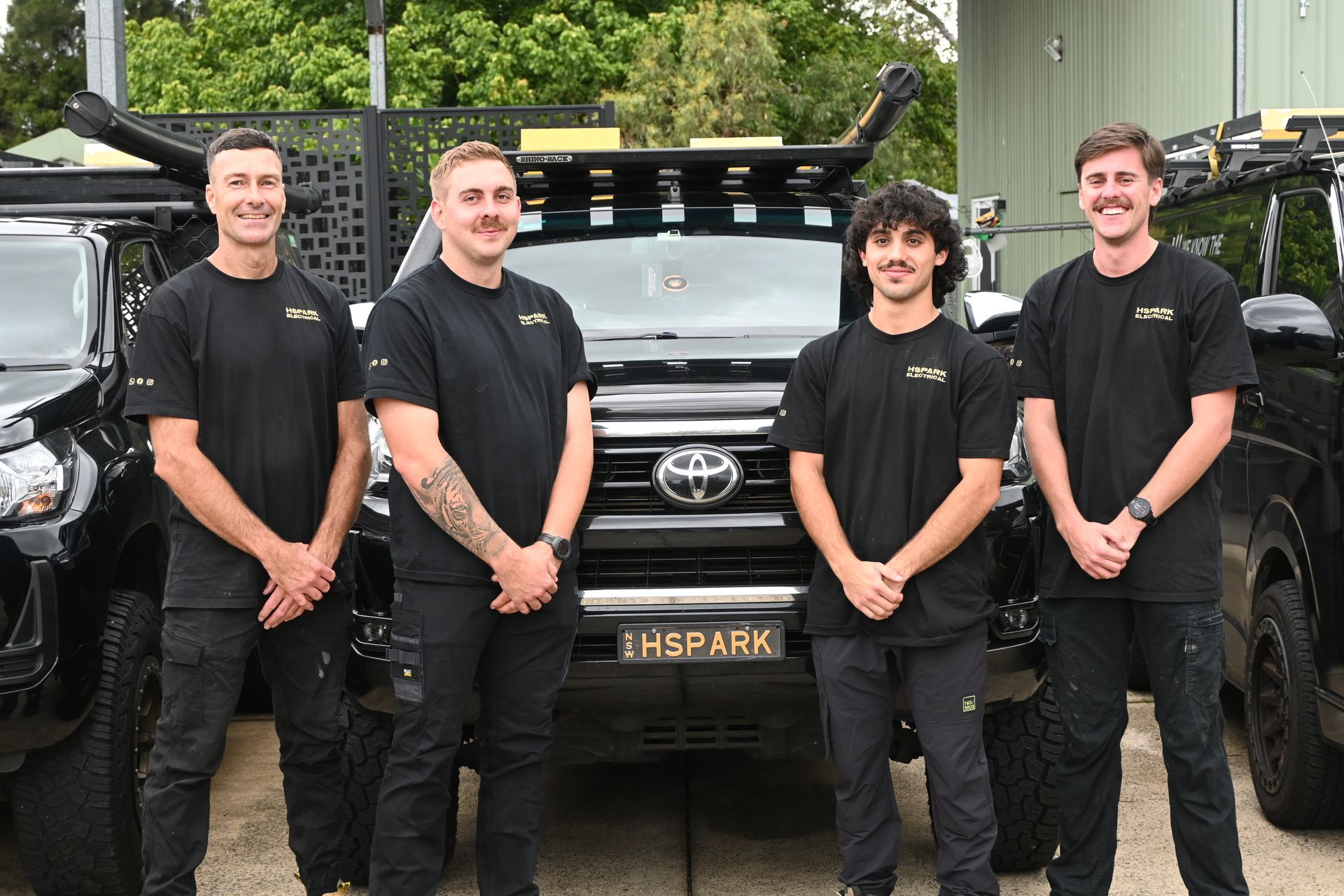 Four smiling individuals wearing matching black shirts stand in front of black vehicles at an outdoor work site — Hspark Electrical PTY LTD in Woonona, NSW