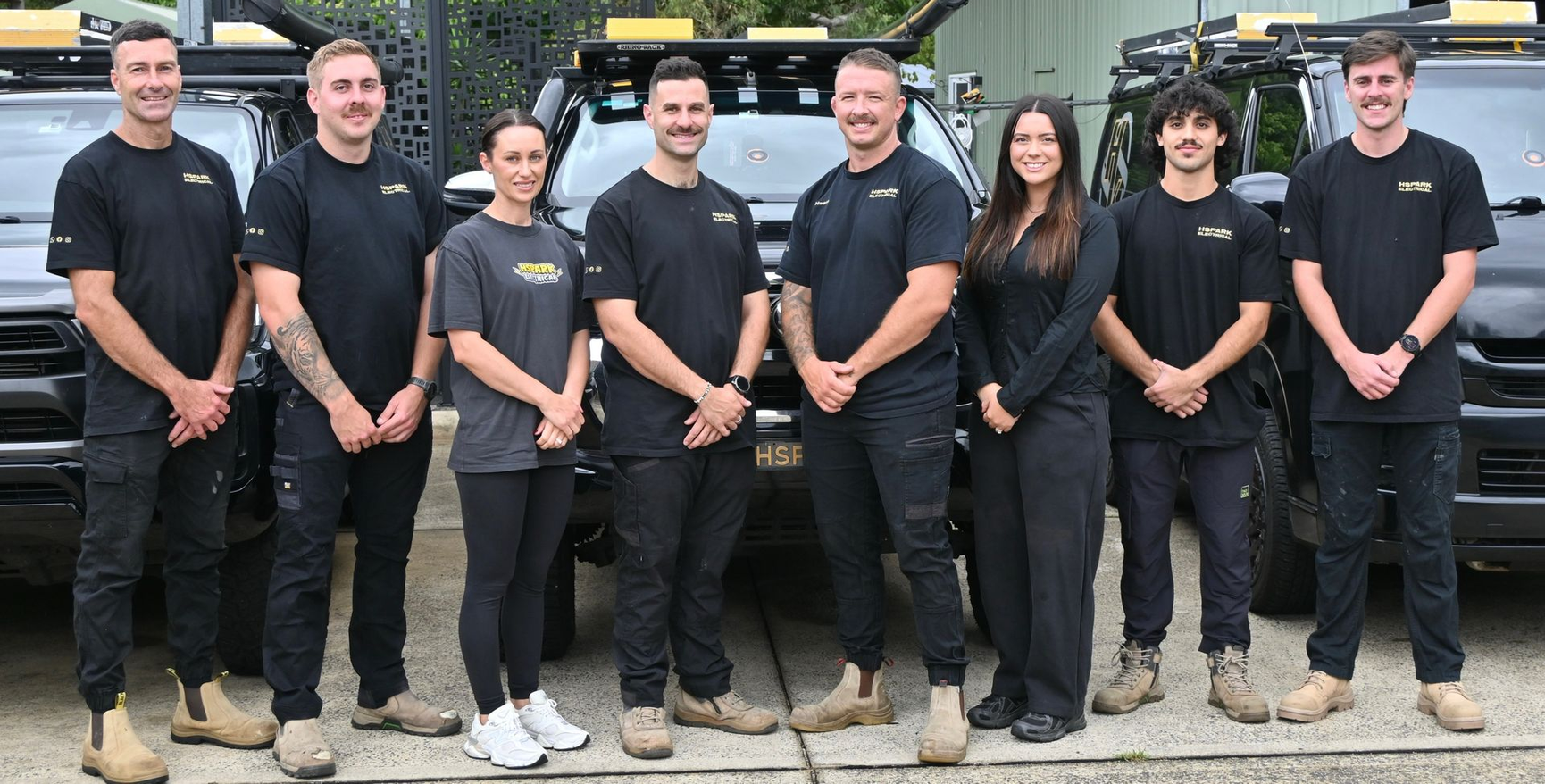 A group of eight people in matching black uniforms stand side-by-side in front of parked utility vehicles — Hspark Electrical PTY LTD in Appin, NSW