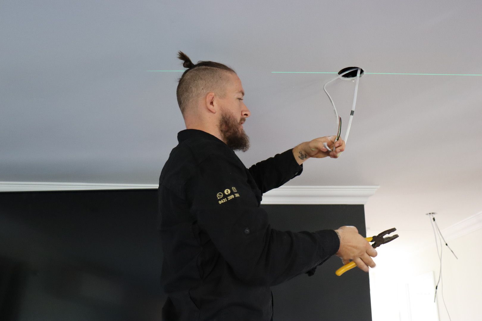 A person with a topknot hairstyle stands on a ladder, using wire strippers to work on ceiling light fixture wiring — Hspark Electrical PTY LTD in Tahmoor, NSW