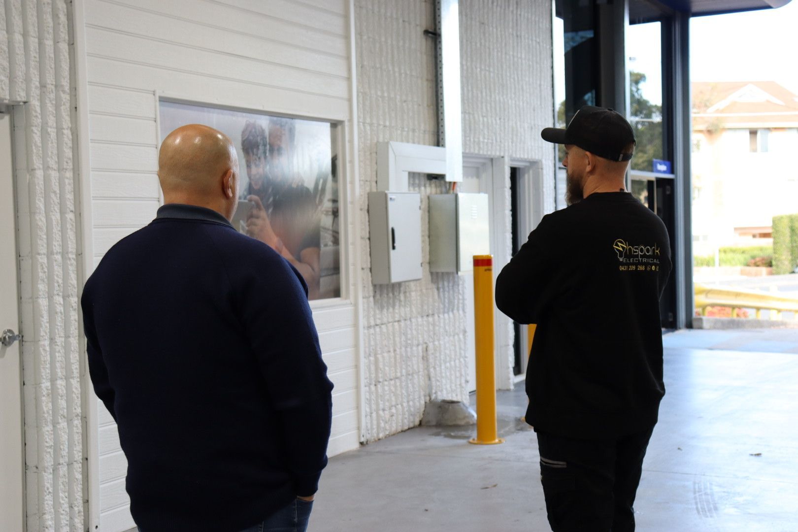 Two people in casual attire stand in an industrial or warehouse space, looking toward a large wall-mounted screen — Hspark Electrical PTY LTD in Appin, NSW
