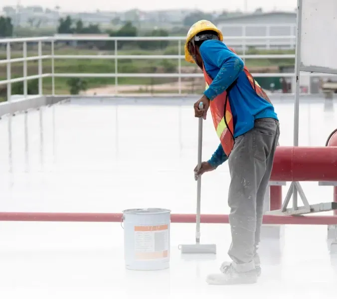 Worker in safety gear painting a rooftop railing with a roller beside a paint bucket.