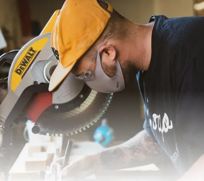 Man in a yellow cap and mask using a circular saw in a workshop, sparks and dust visible