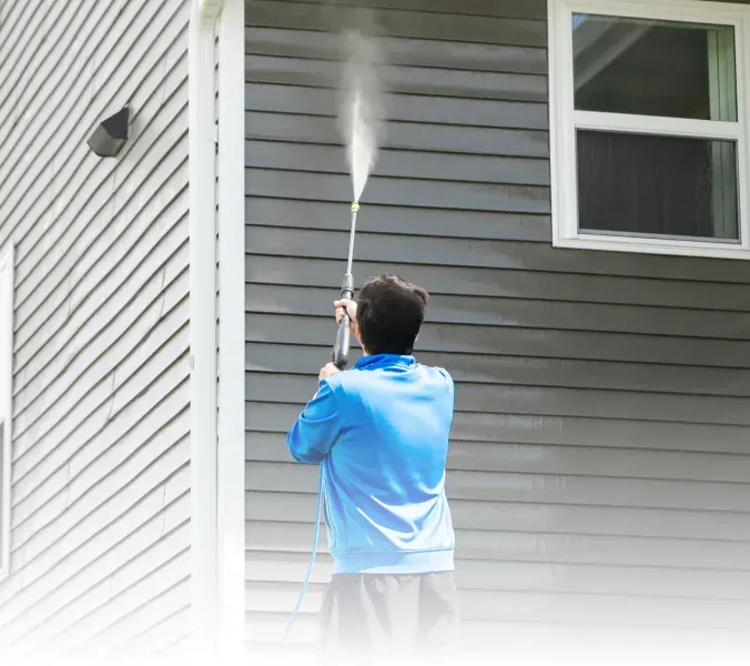 Person power-washing the side of a house with a blue shirt and pressure spray.