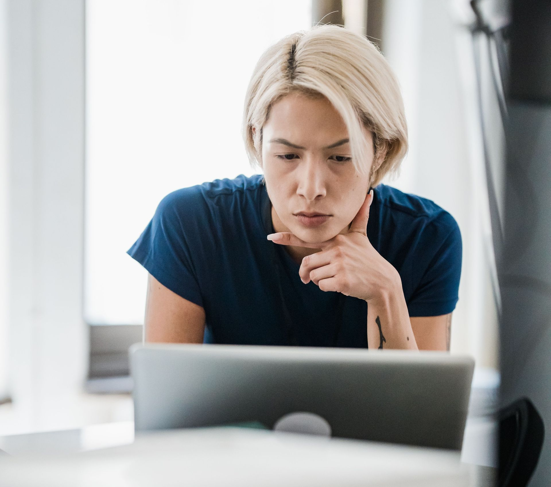 Person with short blond hair studying a laptop at a desk, chin resting on hand, in a bright room