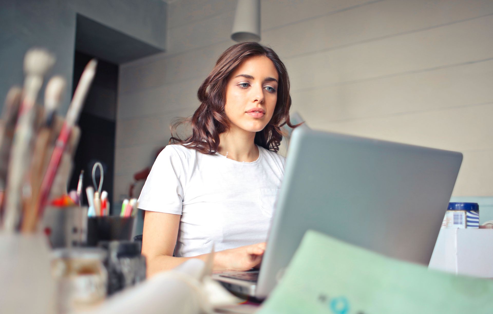 Woman working at a laptop in a bright office, with art supplies blurred in the foreground.