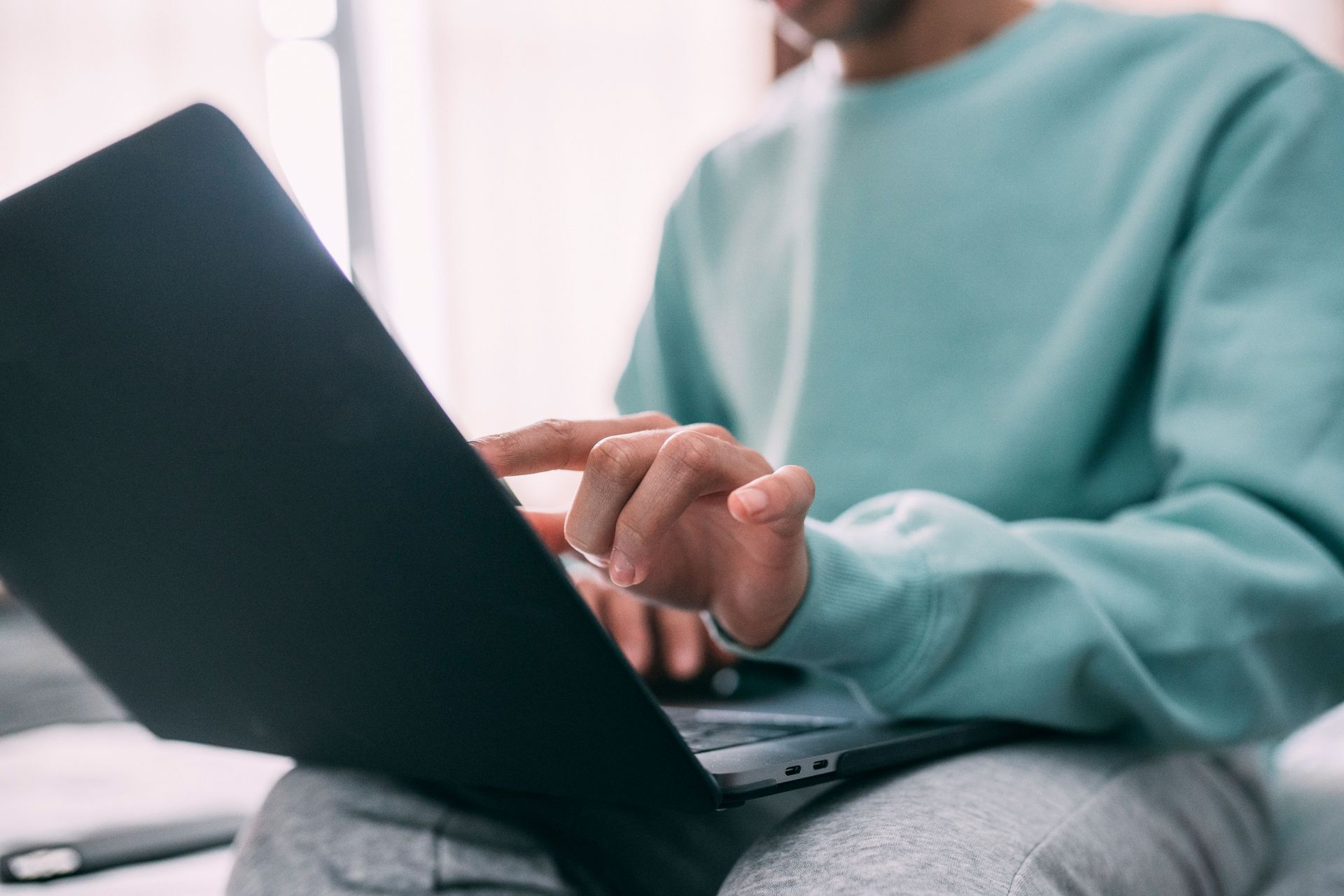 Person using a laptop on a couch, wearing a teal sweater in a bright room