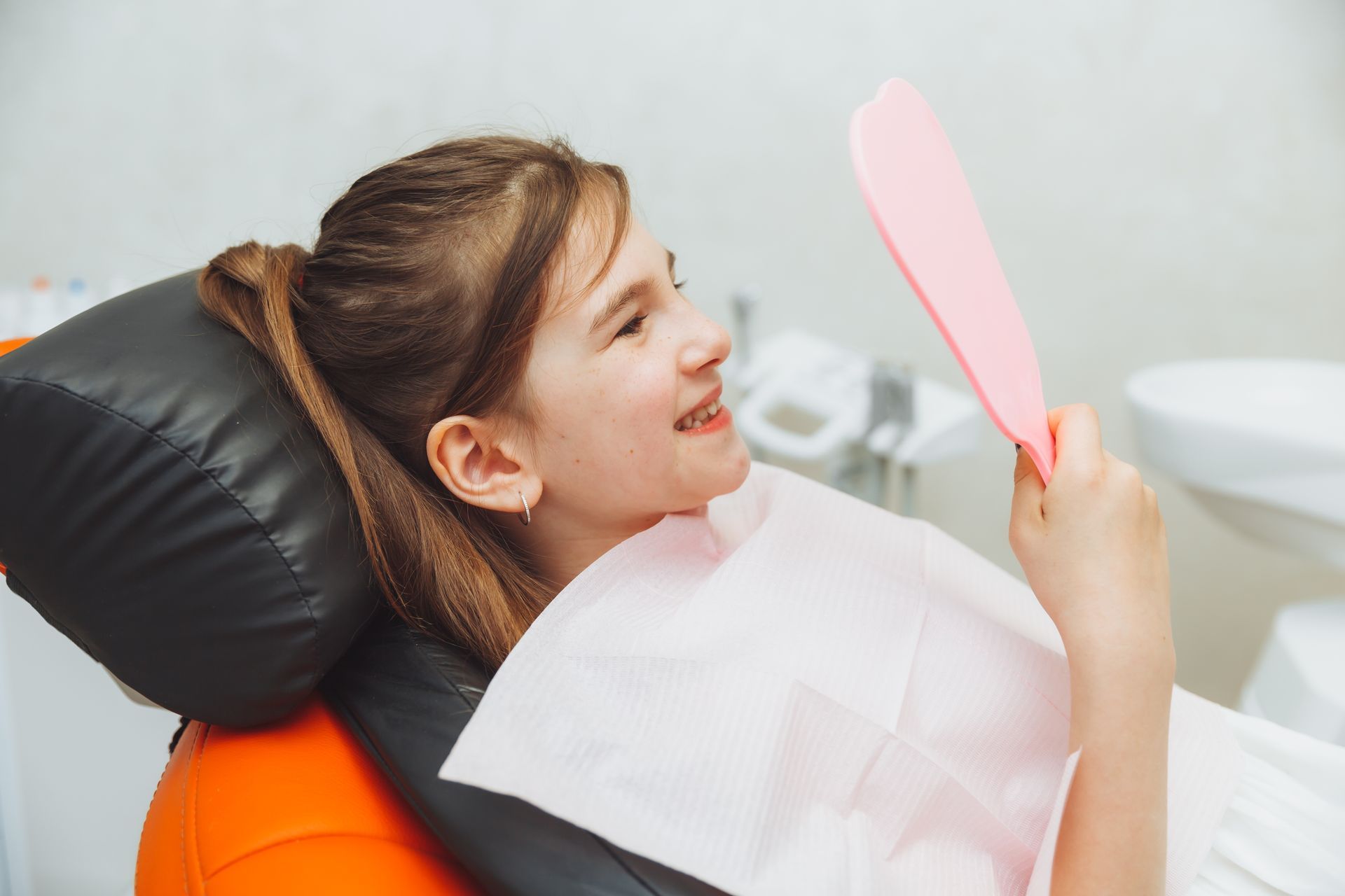 A young girl is sitting in a dental chair looking at her teeth in a mirror waiting for our pediatric dentist in Tarrytown, NY