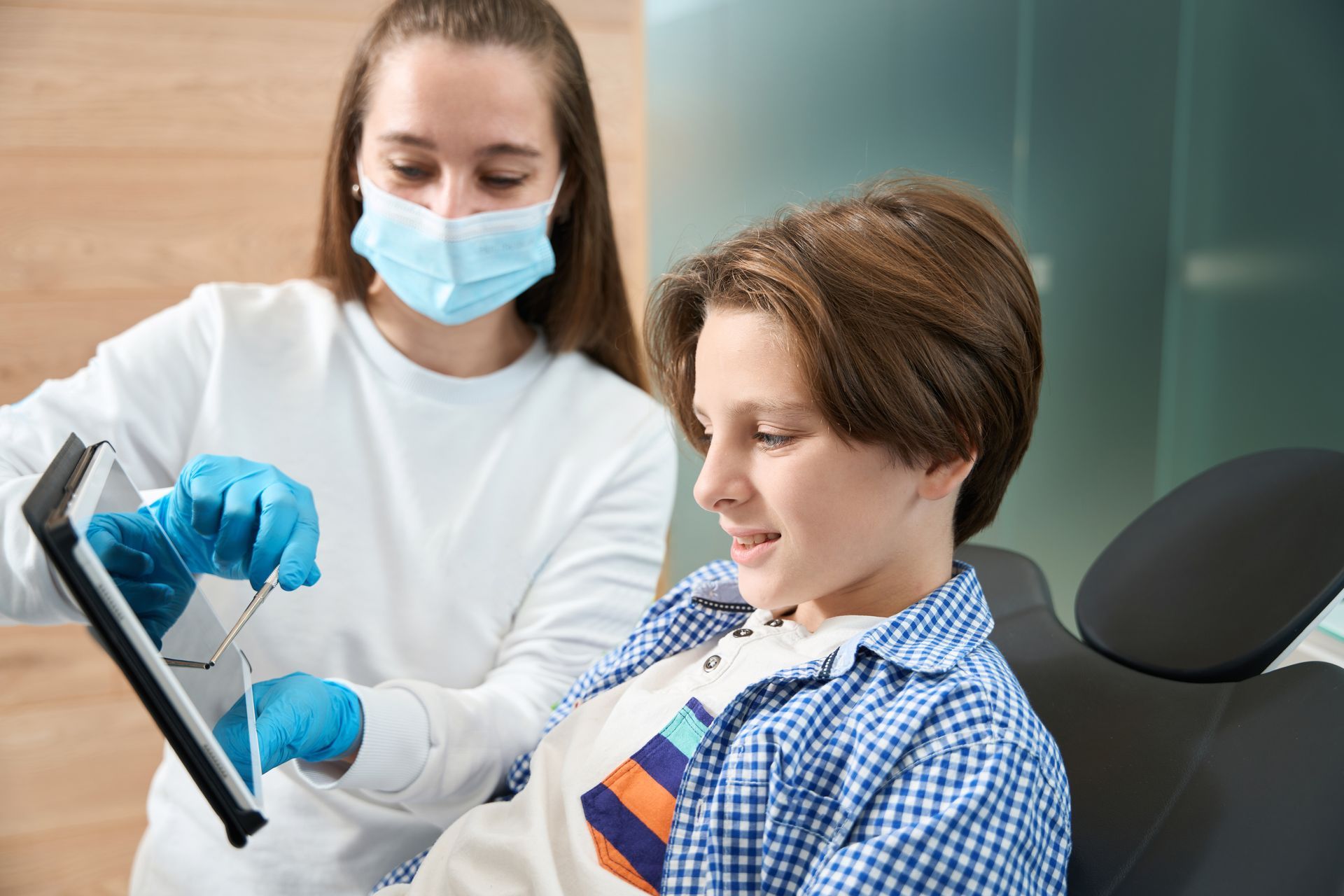 A young boy is sitting in a dental chair while a dentist looks at a tablet.