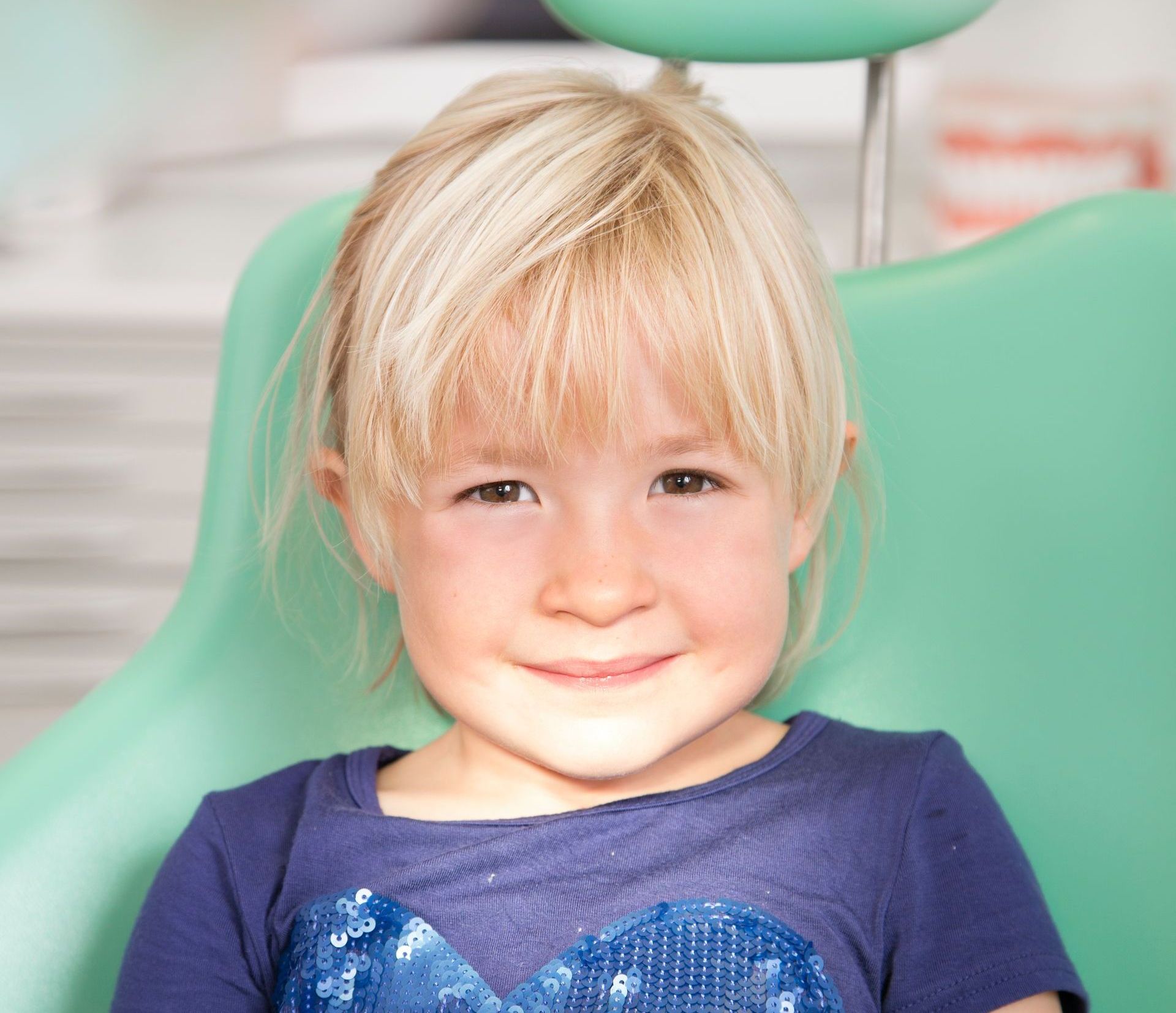 A little girl is sitting in a dental chair and smiling.