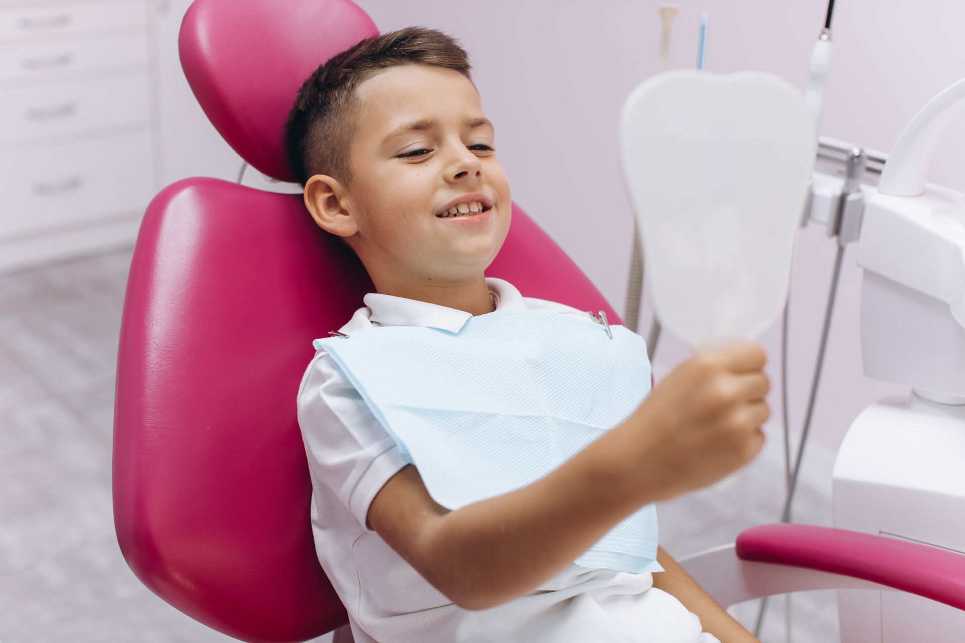 A young boy is sitting in a dental chair looking at his teeth in a mirror.
