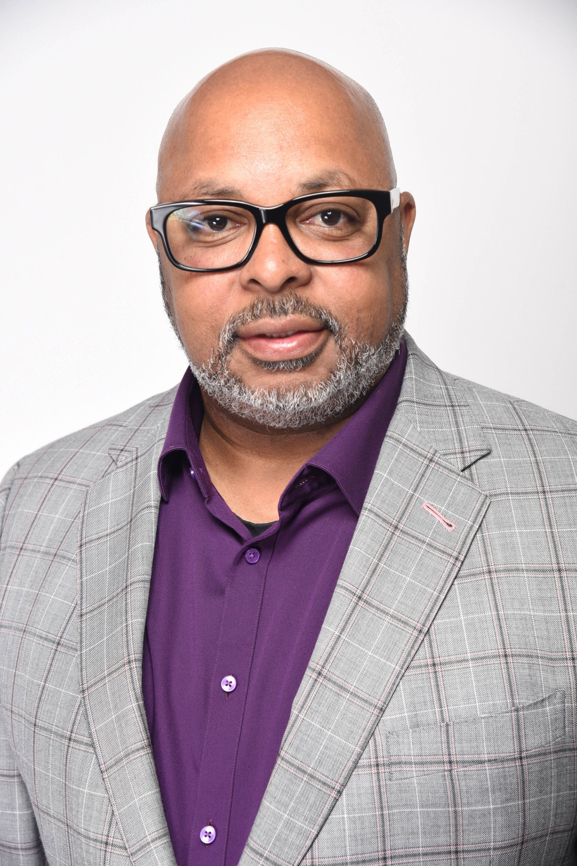 A bald man wearing glasses and a purple shirt is standing in front of a white background.