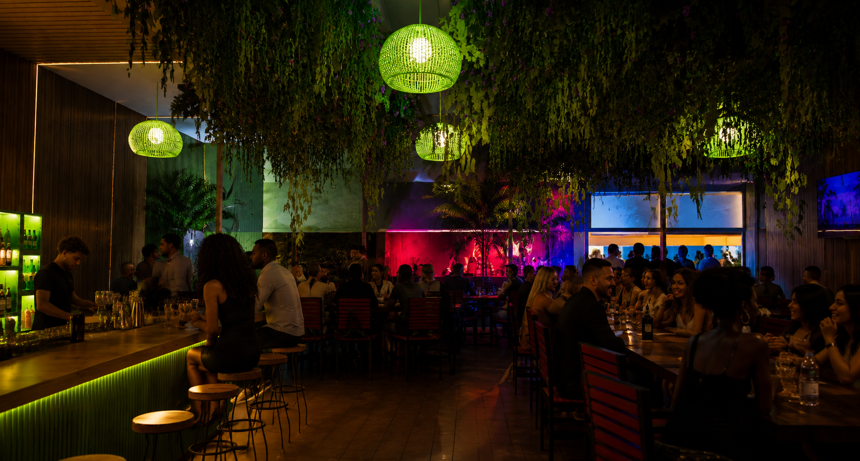 Dimly lit tropical bar with hanging greenery, green pendant lights, and people seated at tables.