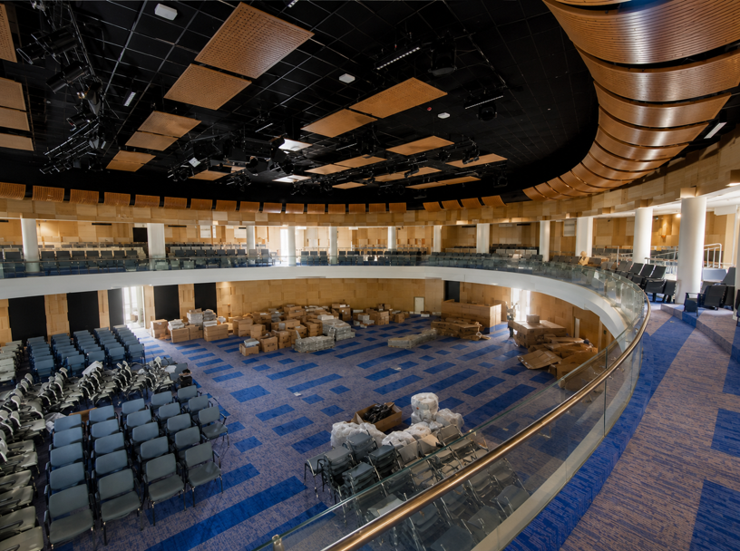 Empty indoor auditorium with blue seating, curved balcony, and exposed ceiling panels under renovation