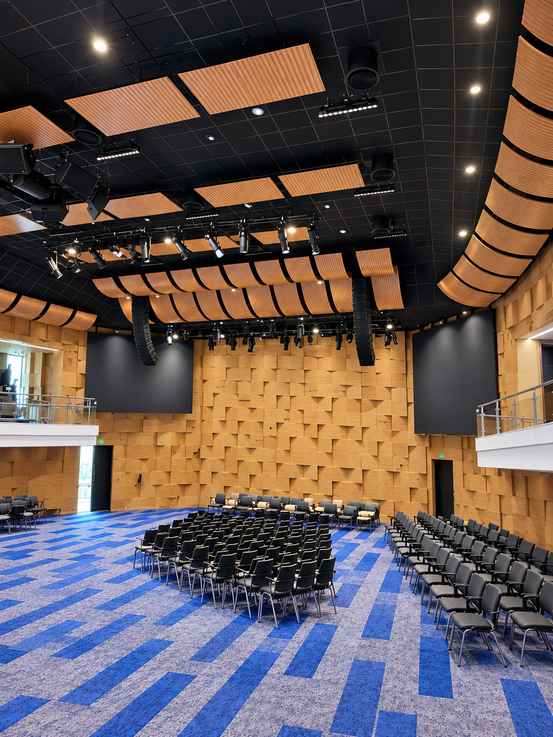 Empty auditorium with blue patterned carpet, black seats, and wood acoustic wall panels under a dark ceiling.