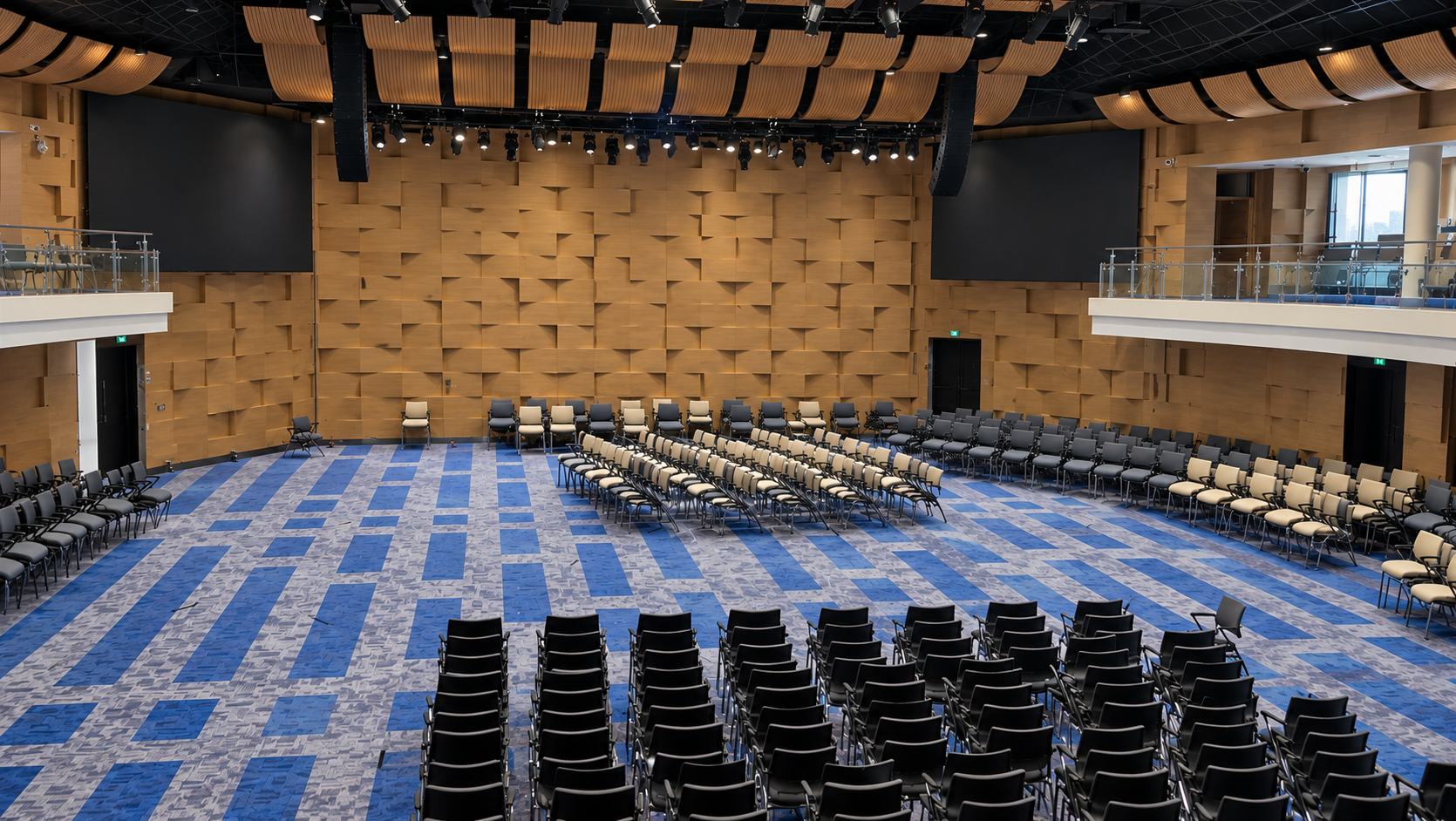 Empty auditorium with rows of black chairs and a blue carpeted floor facing a tan stage wall.