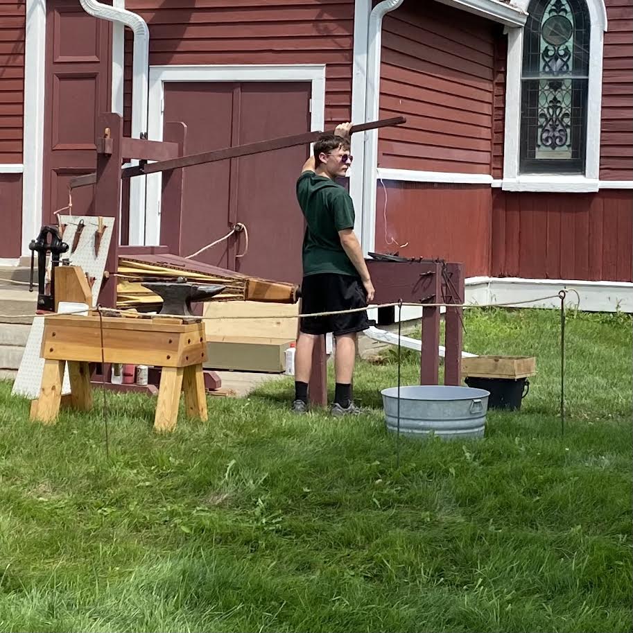 Person working at a blacksmith setup outside a red building, near a water basin and grass.