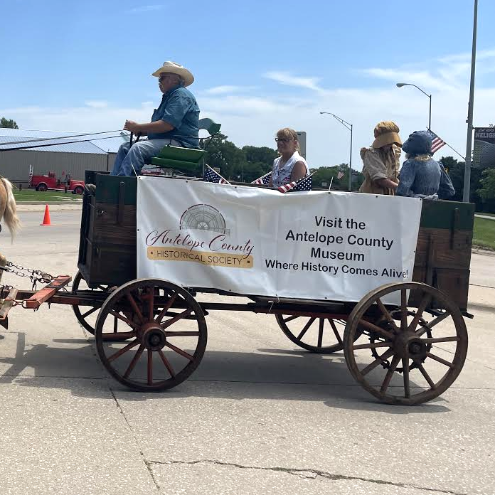 Horse-drawn wagon with people, banner: Antelope County Museum. In a parade, outdoor setting.
