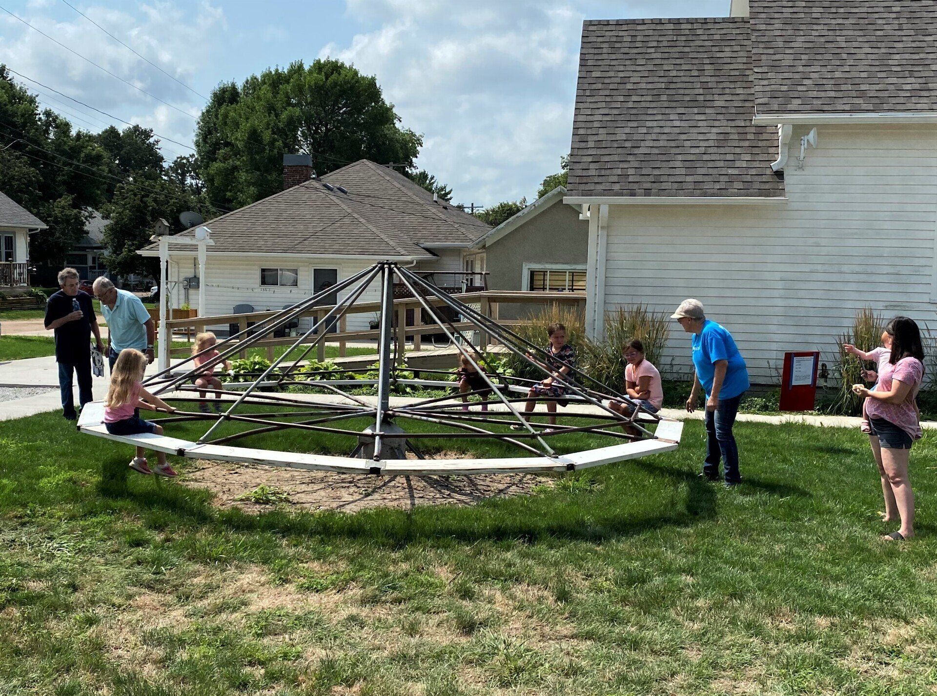 Children on a vintage metal merry-go-round, with adults nearby, in a grassy yard, near white buildings.