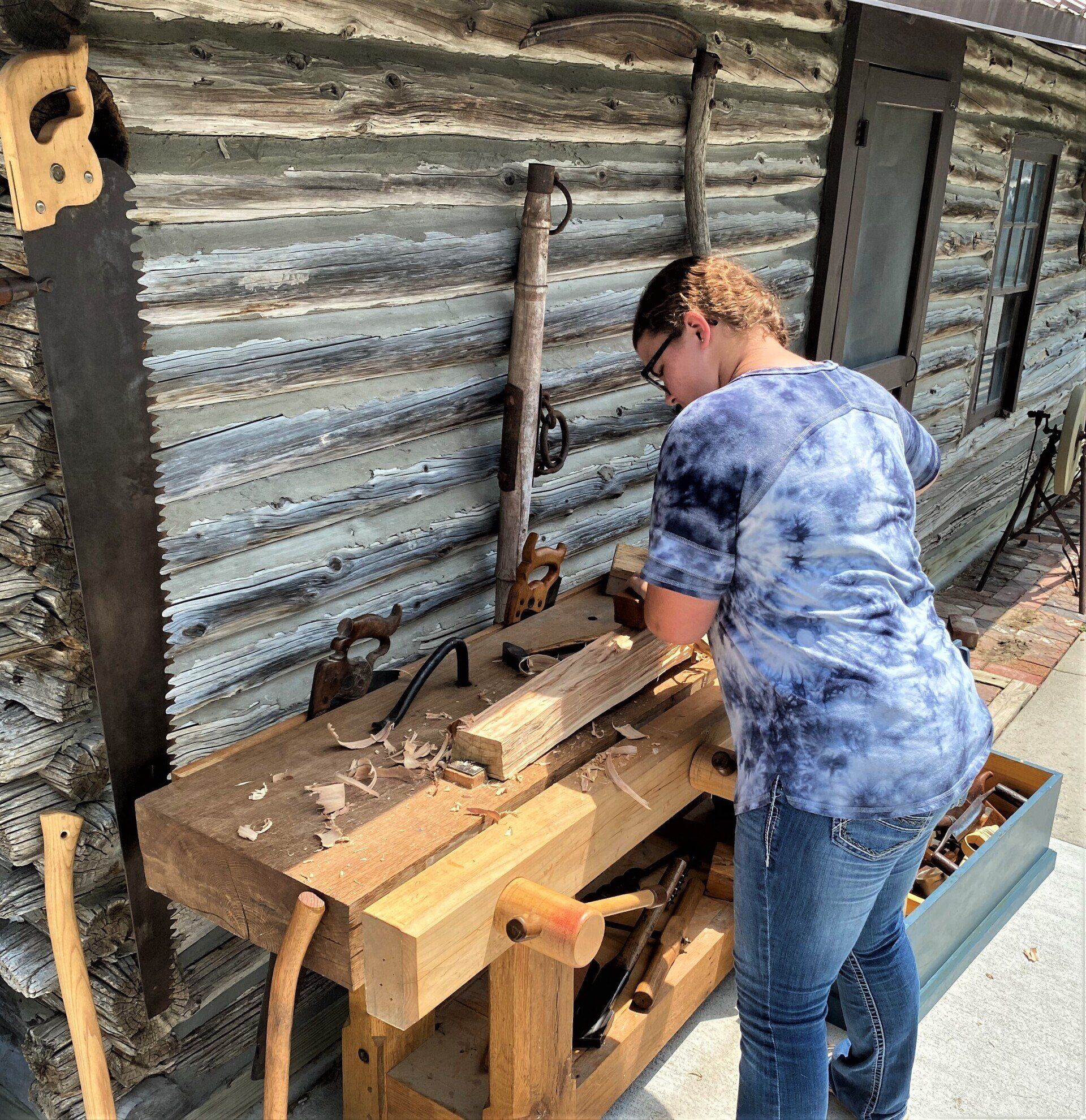 Person using hand tools at a wooden workbench. Outdoor setting with tools leaning against a log cabin.