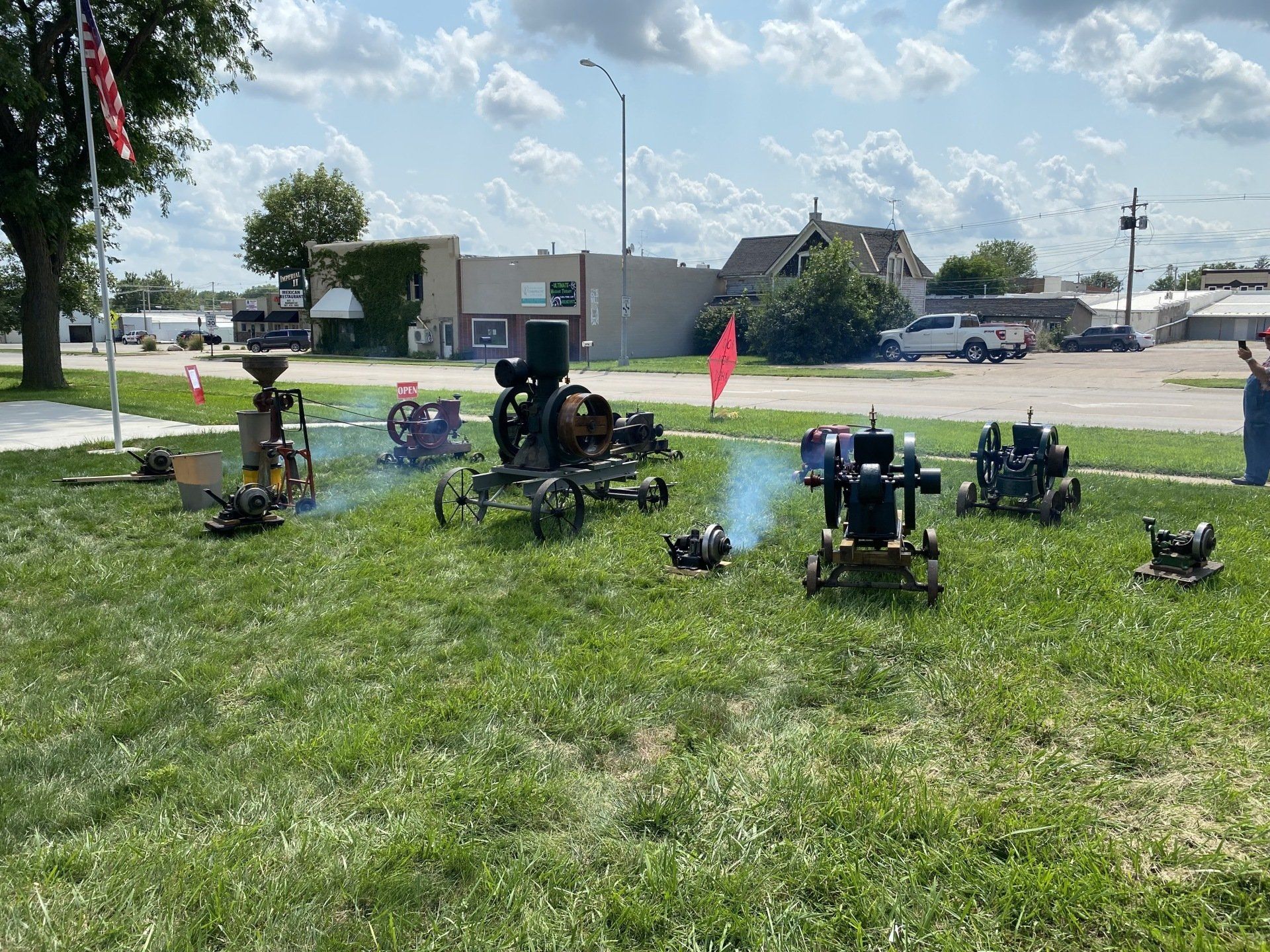 Antique engines on grass display, emitting smoke. Buildings and a street in background, sunny day.