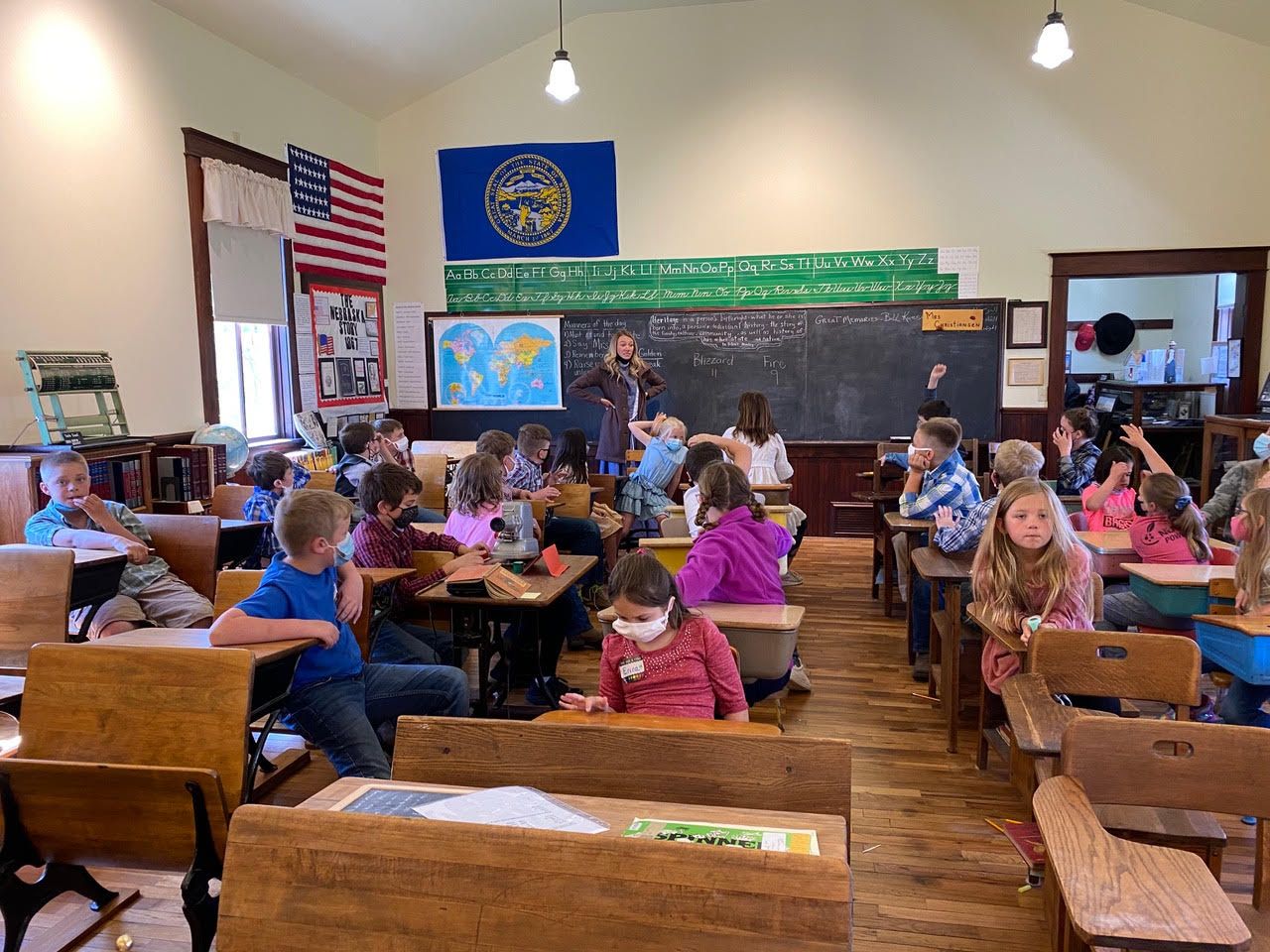 Children in an old classroom, with a teacher. Some students raise hands. American flag and world map on the wall.