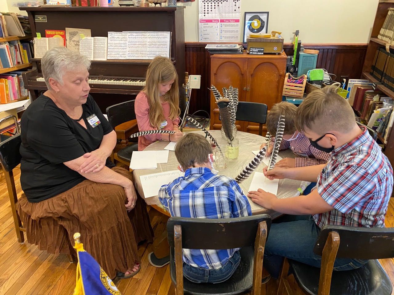 Children at a round table writing with quill pens; an adult watches; library setting.