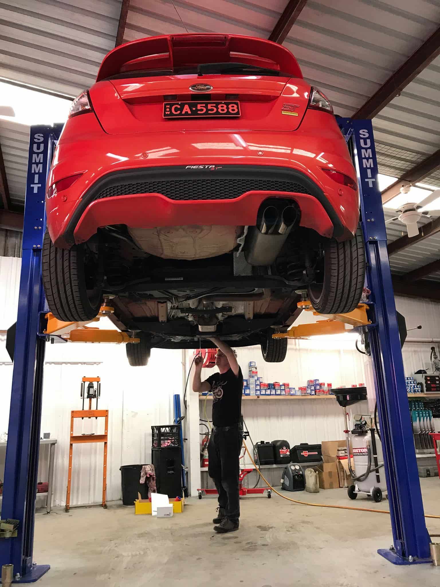 A Man Is Working Under A Red Car On A Lift In A Garage — Sojos Mechanical & Tyre Service In West Gosford, NSW