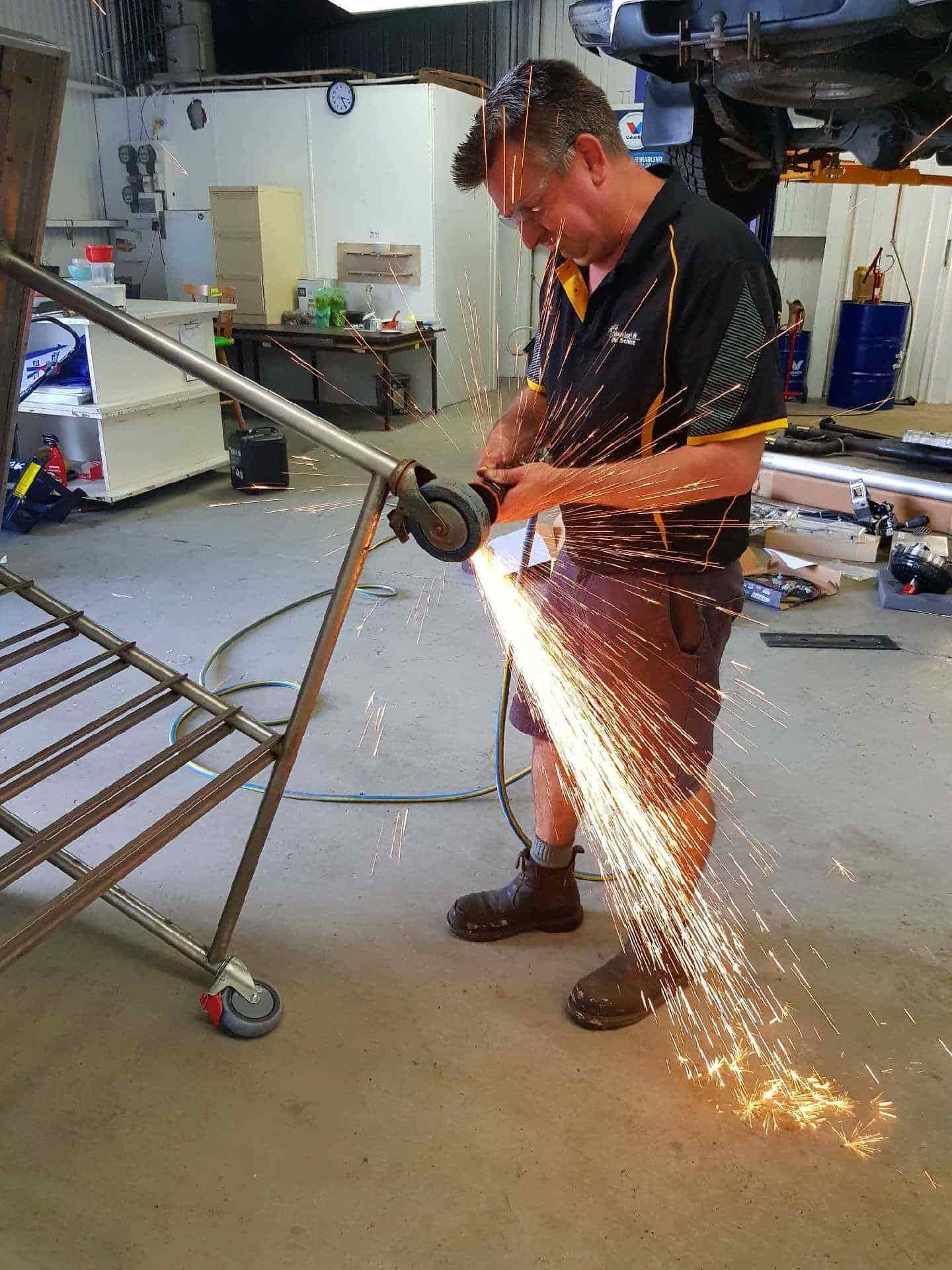 A Man Is Cutting A Metal Pipe With A Grinder In A Garage — Sojos Mechanical & Tyre Service In Kariong, NSW