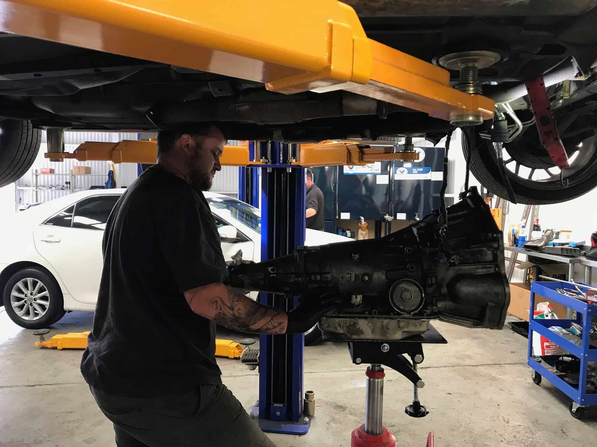 A Man Is Working Under A Car On A Lift In A Garage — Sojos Mechanical & Tyre Service In West Gosford, NSW