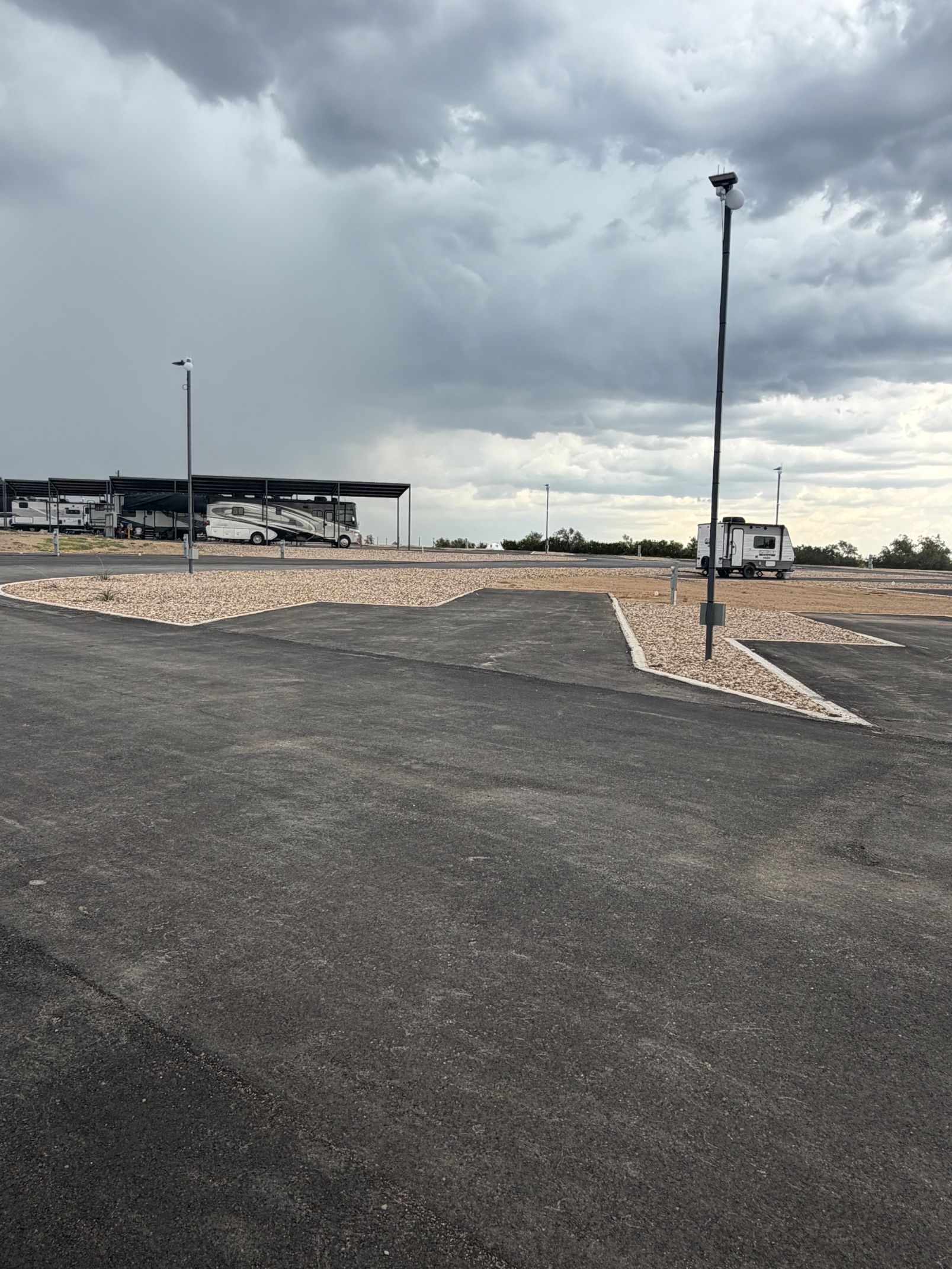 Empty paved lot under cloudy sky with light poles and a covered structure in the distance