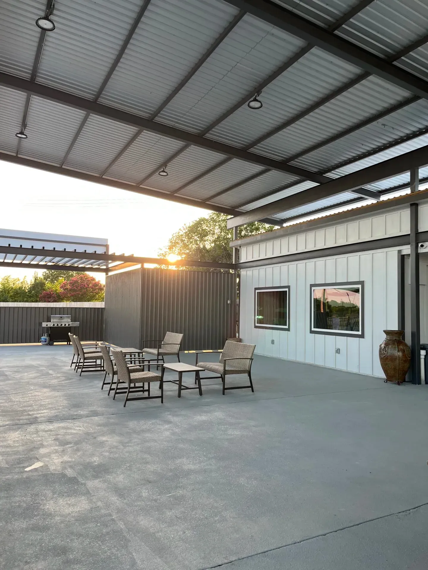 Covered patio with seating, string lights, and sunset view beside a white building and black container wall.