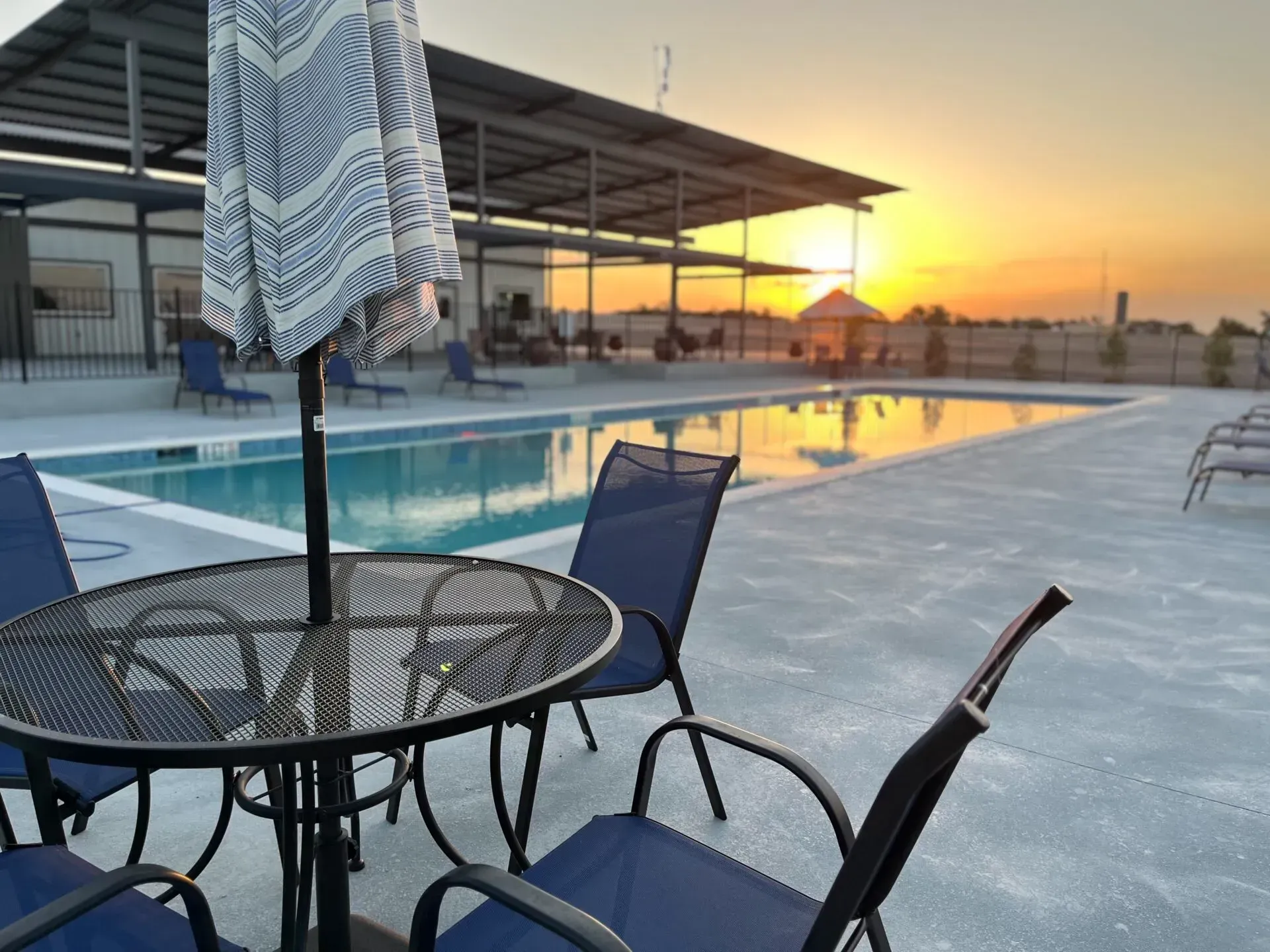 Outdoor pool deck at sunset with patio table, chairs, and an umbrella beside the water