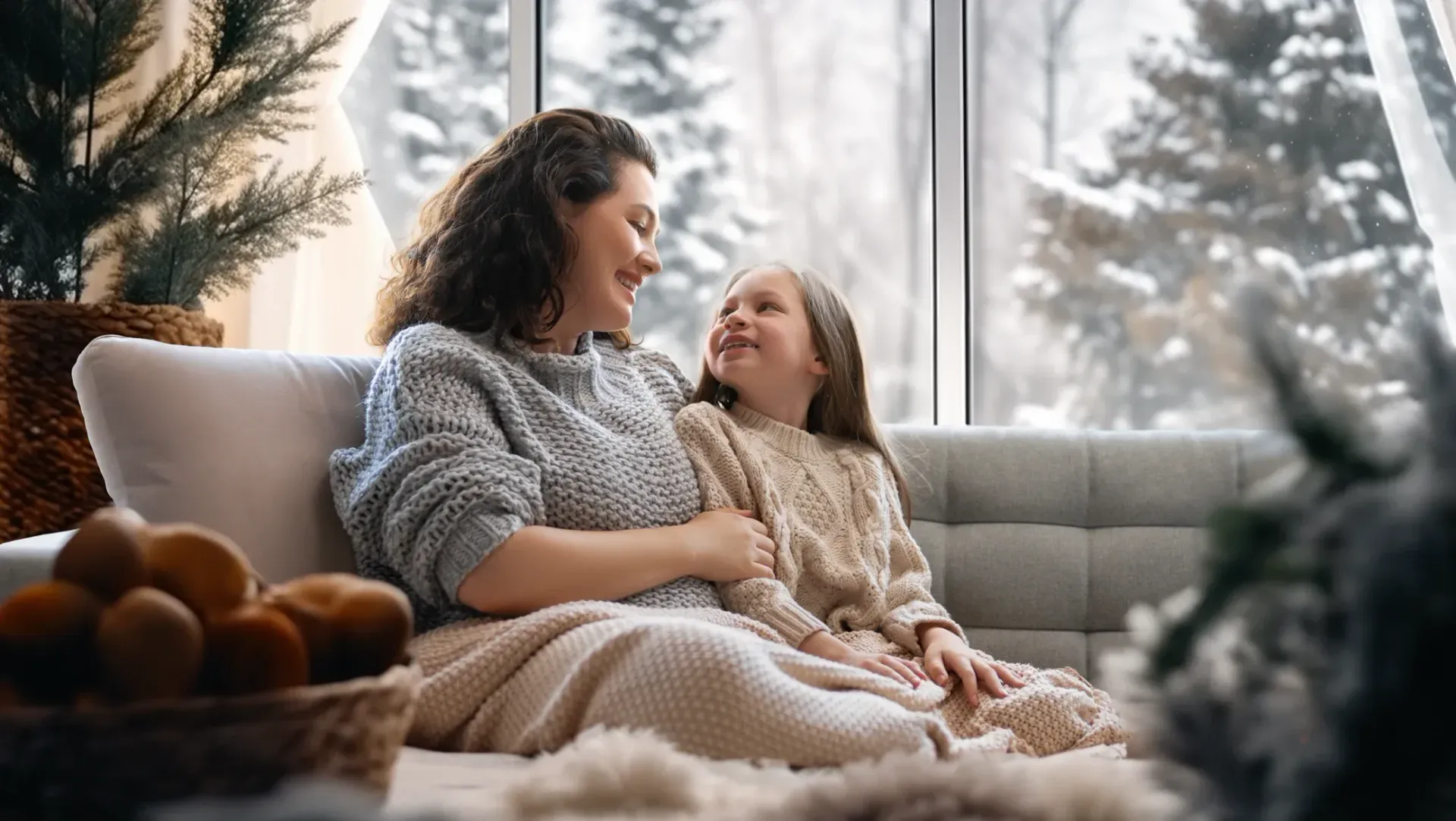 Woman and child, smiling, sitting on a sofa by a window overlooking snow-covered trees.