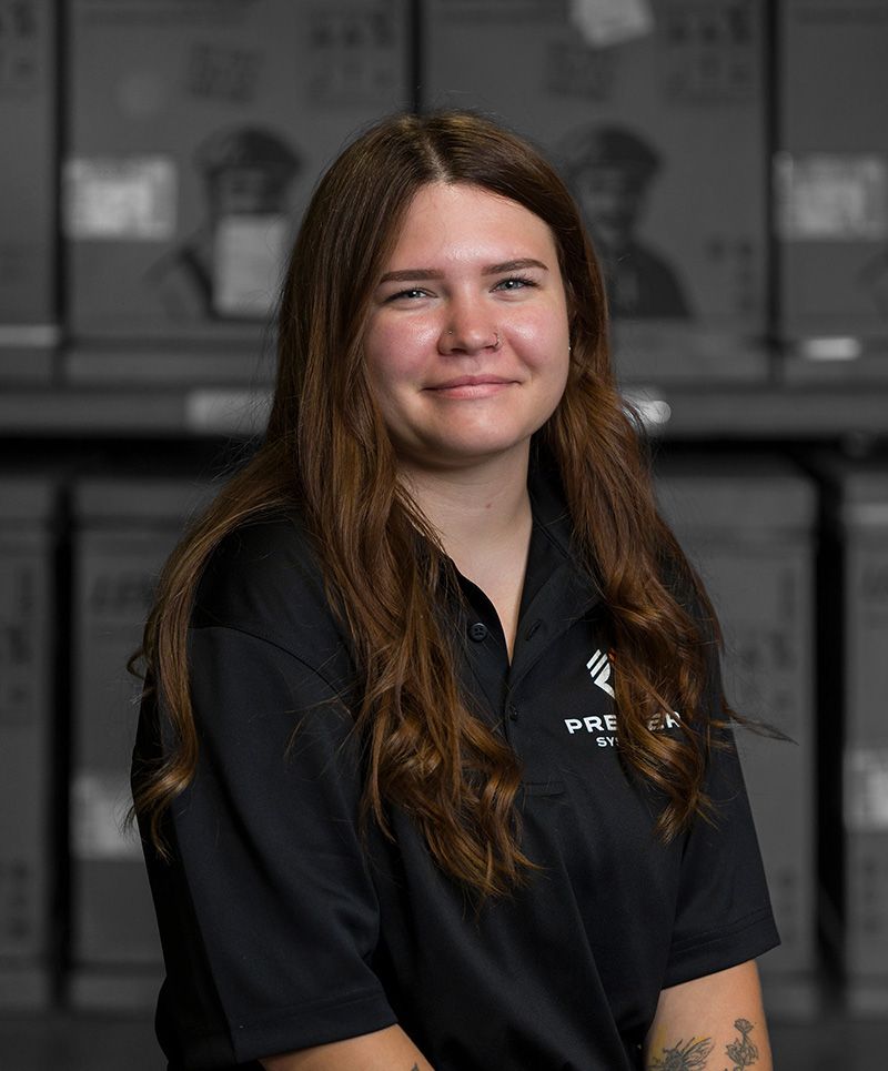 Woman in black polo shirt smiles, posing in front of boxes, likely a warehouse setting.