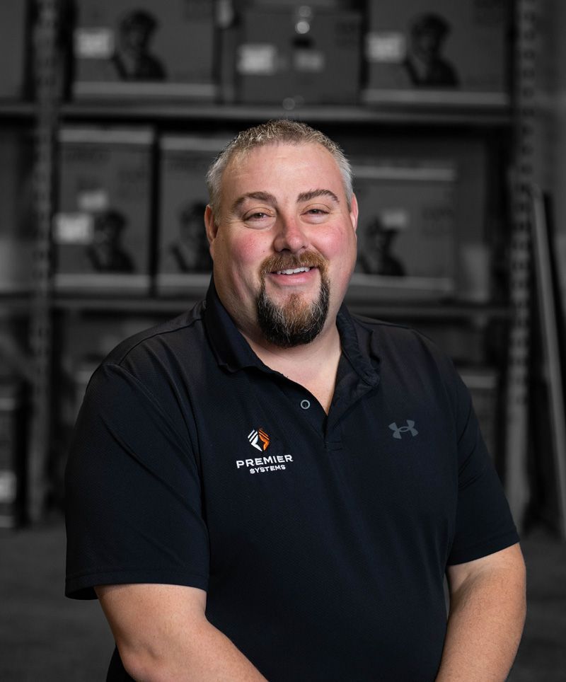 Man wearing a black polo shirt, smiling. Warehouse shelves are in the background.