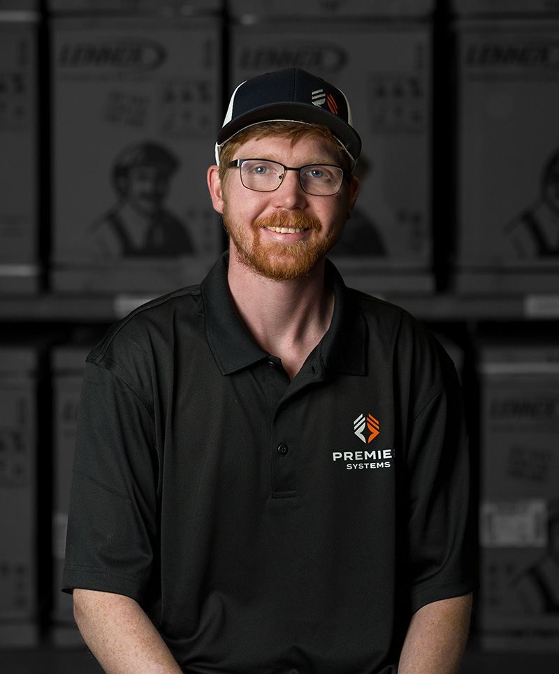 Man with red hair and glasses wearing a black polo shirt and baseball cap smiles in front of storage boxes.