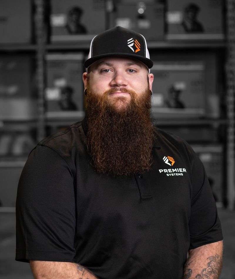 Man with beard wearing a cap and black shirt with a company logo, in a warehouse.