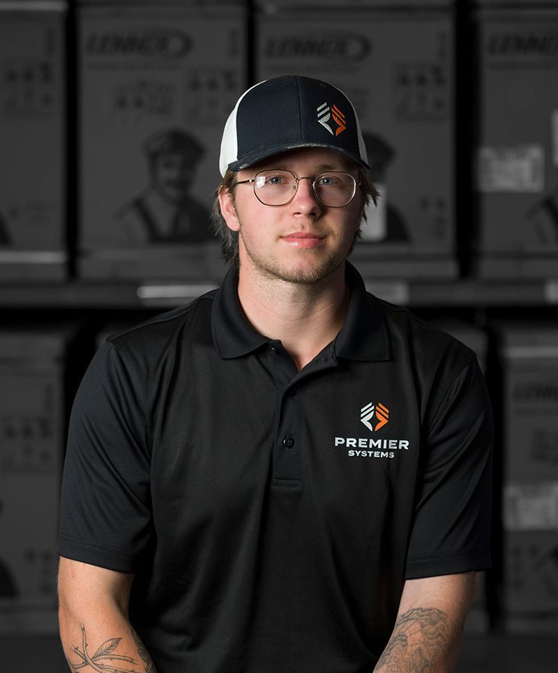 Man in black polo and cap, glasses, arms crossed, in warehouse setting with boxes.