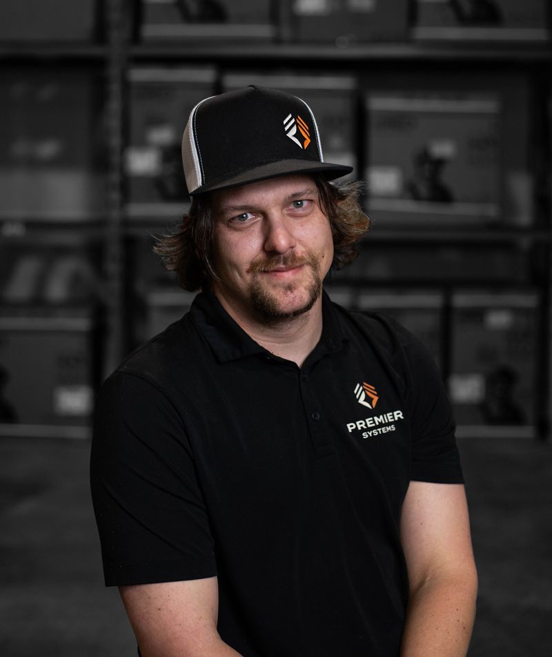 Man in black polo shirt and cap smiling, logo visible; warehouse background.
