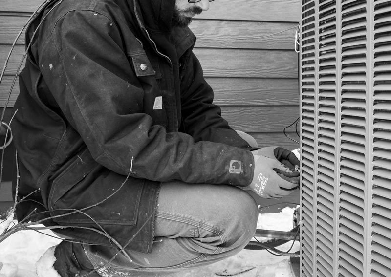 Person wearing work clothes, kneeling near an outdoor air conditioning unit. Hands gloved, performing maintenance in a snowy setting.