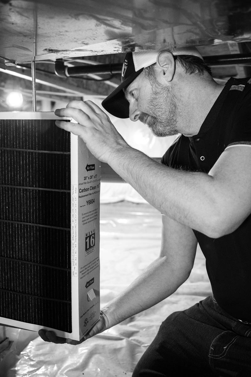Man in cap replacing air filter, holding it up near ductwork.