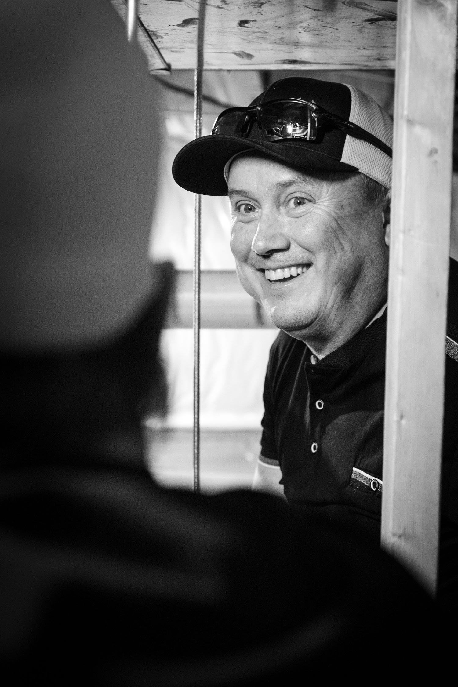 Man wearing a hat smiles, looking toward another person standing nearby. Black and white photo.